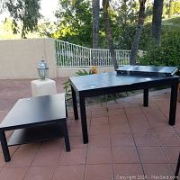 Black Ikea dining table with two leaves positioned on top and black Ikea sofa table against outdoor backdrop on tiled patio.