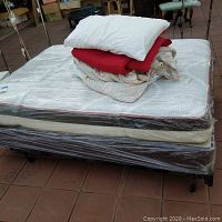 Full bedframe and mattress wrapped in protective plastic with stacked linens including pillow, sheet, and two comforters