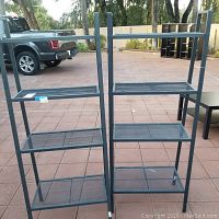 Front view of two metal mesh shelf units side by side outdoors on tile ground near outdoor furniture and truck.