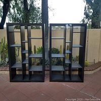 Front view of two matching black wood shelving units with geometric shelving design outdoors on tiled floor.