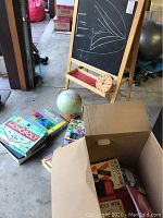 Wooden foldable chalkboard with black chalk surface, attached wooden clock, globe, Monopoly board game, Super Mario chess set, and box of educational books visible