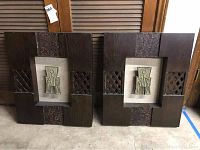 Two heavy wood wall art frames leaning against wooden shuttered doors, showing dark wood with carved lattice and embossed detail sections, each with central beige fabric inset and abstract metal figure.
