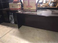 Large dark-colored desk with solid panels, showing surface scratches and marks, placed on a concrete floor with cardboard boxes on top.
