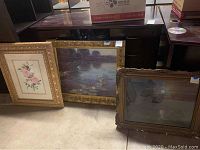 Three framed paintings leaning against a dark piece of furniture showing their fronts and frames. Two framed artworks have gold ornate frames and one frame is visibly worn. Included floral and landscape paintings.