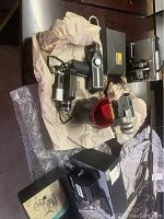 Photo showing various vintage movie cameras and projectors arranged on a table with bubble wrap and paper padding. Several cameras visible including one with a lens attached, black and silver metal/plastic construction. Projectors nearby also visible.