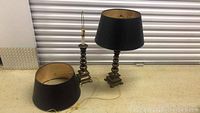 Two brass lamps with black shades, one lamp assembled, one lamp is missing its shade on top