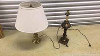 Two brass table lamps side by side, one with a white shade and one without shade, showing electric cords and plugs on a concrete floor.
