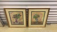 Front view of two framed paintings showing colorful floral arrangements in vases with artist signatures.