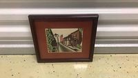 Front view of the framed cross-stitch main street artwork leaning against a white wall.