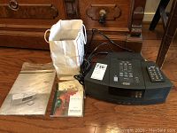 Bose Wave Radio/CD Player with power cord, remote control, user manual, white gift bag, and a couple of CDs on a wooden floor.