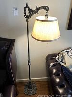 Full view of vintage pewter color base floor lamp with light-colored lampshade lit up and standing next to furniture.