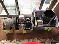 Three boxes filled with various kitchenware items including pots, pans, bowls, and colander, stacked by a window.