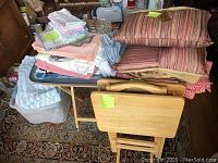 Lot composed of linens and pillows stacked on and around folding tray tables.