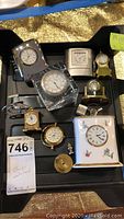 Photo showing nine assorted clocks arranged in a black tray, including gold-tone, silver-tone, crystal, and ceramic models.