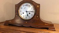 Full front view of vintage mantle clock showing shape, dial, and wooden case with decorative inlay.