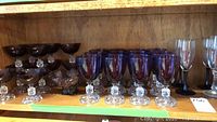 Wide view of the set showing purple champagne glasses, water glasses, and four clear champagne flutes with black stems arranged on wooden shelf.
