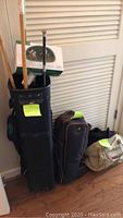 Photo of the black nylon golf bag standing upright with wooden polo mallets inside, along with other bags on the floor nearby.