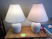 Pair of two cream ceramic lamps with ribbed bases and white fabric shades on wooden surface, both lamps lit, paint marks visible on one shade