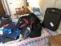 Photo showing assorted bags including black-red duffle bag, blue tote bags, and black soft bag on a bed next to a chair and table.