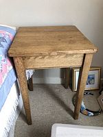 Square small side table with visible wood grain and slightly worn natural wood finish placed beside a bed.