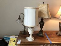 Three table lamps on a wooden desk: white marble base lamp with white fabric shade, brass lamp with two arms and glass shades, brass/bronze lamps with ornate decorative shades with tassels.