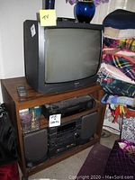 Admiral 19 inch CRT TV on wooden shelf with remote on top shelf, Sharp VHS player in middle compartment, AIWA stereo system with two speakers in bottom compartment beside folded fabric.