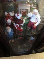 Multiple Christmas Santa figurines arranged in cabinet including small Santa in blue jacket, traditional Santa figures, and Christmas fireplace decoration
