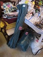 View of stacked linens, including drapes, folded fabric curtains, and linens with assorted colors. Also visible silverware and glassware on table.