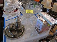 Full lot view showing punch bowl, silver plated tea service on tray, Baby Cakes mini cupcake maker in box, and linens in background.