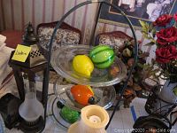 Overview showing decorative glass fruit in clear glass bowls on metal serving stand with some lanterns and floral arrangement in background.