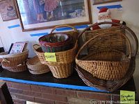 Photo showing a collection of various woven baskets of different sizes and styles stacked and arranged on a brick ledge, including a basket labeled 'baskets' and a glimpse of recipe books behind them.