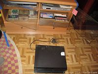 Photo showing black Shintom VCR player on the floor beneath a cabinet containing VHS tapes and DVD player.