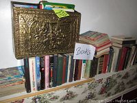 Books of various genres arranged on a shelf with a gold-colored metal embossed magazine holder placed on top of the shelf.