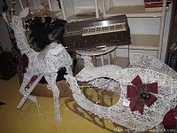 White woven outdoor Christmas reindeer decoration with string lights and some legs detached or needing reattachment, next to a similar white woven sleigh decoration with red bow.