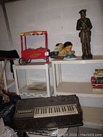 Wide view showing the soldier statue, boy with frog statue, red plastic hot dog maker, shelving and vintage organ below