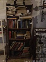 Lighted photo showing the entire brown MDF bookcase with four shelves filled with numerous books in a basement or storage area with concrete walls.