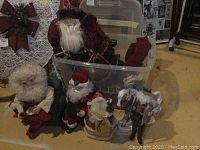Multiple Santa figurines placed around a plastic bin with lid on a floor, including a red velvet Santa and a white-haired Santa holding money, and a paper horse figurine.