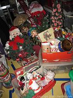 Various Christmas figurines, plush toys, wreaths, tins, decorative ladder and other holiday decor items packed together in a box and on table surfaces.