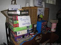 Stack of puzzles and board games including 1000 and 2500 piece puzzle boxes, Trivial Pursuit and Scrabble boxed games on top of a chair.