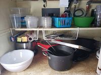 Photo of kitchen shelf with white mixing bowl, large black pot with lid and handles, assorted plastic containers in blue and green baskets, and other containers.