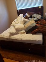 Full view of queen bed with wooden frame, headboard, mattress, pillows, and linens piled on top. Bed includes a drawer in the footboard for storage.