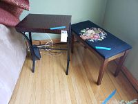 Side view of the vintage side table and needlepoint piano bench on hardwood floor near a wall, highlighting table finish and bench upholstery