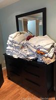 Front view of dresser with stacks of towels and linens on top. Shows dark wood dresser with six drawers and attached mirror.