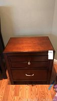 Front view of a dark brown wooden nightstand with two drawers and a small metal knob on the top drawer and a horizontal handle on the bottom drawer.