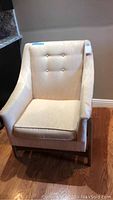 Upholstered armchair with button tufted backrest and cushioned seat, beige fabric, wooden legs on hardwood floor.