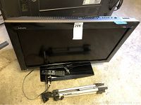 Full image of Sharp Aquos TV with remote resting on its base and Kodak tripod lying next to it on floor.