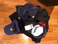 Basket of men's clothing items featuring four baseball caps on top with black, blue with crown logo, and two white and red/blue Cabela's caps. Underneath are folded dark colored North Face jackets and a pair of dark jeans.