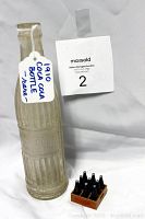 Antique Coca Cola bottle with tag indicating year 1910 and mention of condition, next to numbered auction tag
