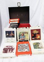 Open brown vinyl case with 8 eight-track tapes and 6 cassette tapes inside, showing album art of various artists including John Denver and Chicago