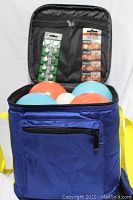 Blue nylon carry bag opened showing four bocce balls (orange and blue) and one white pallino ball with two types of button cell batteries inside interior compartments.